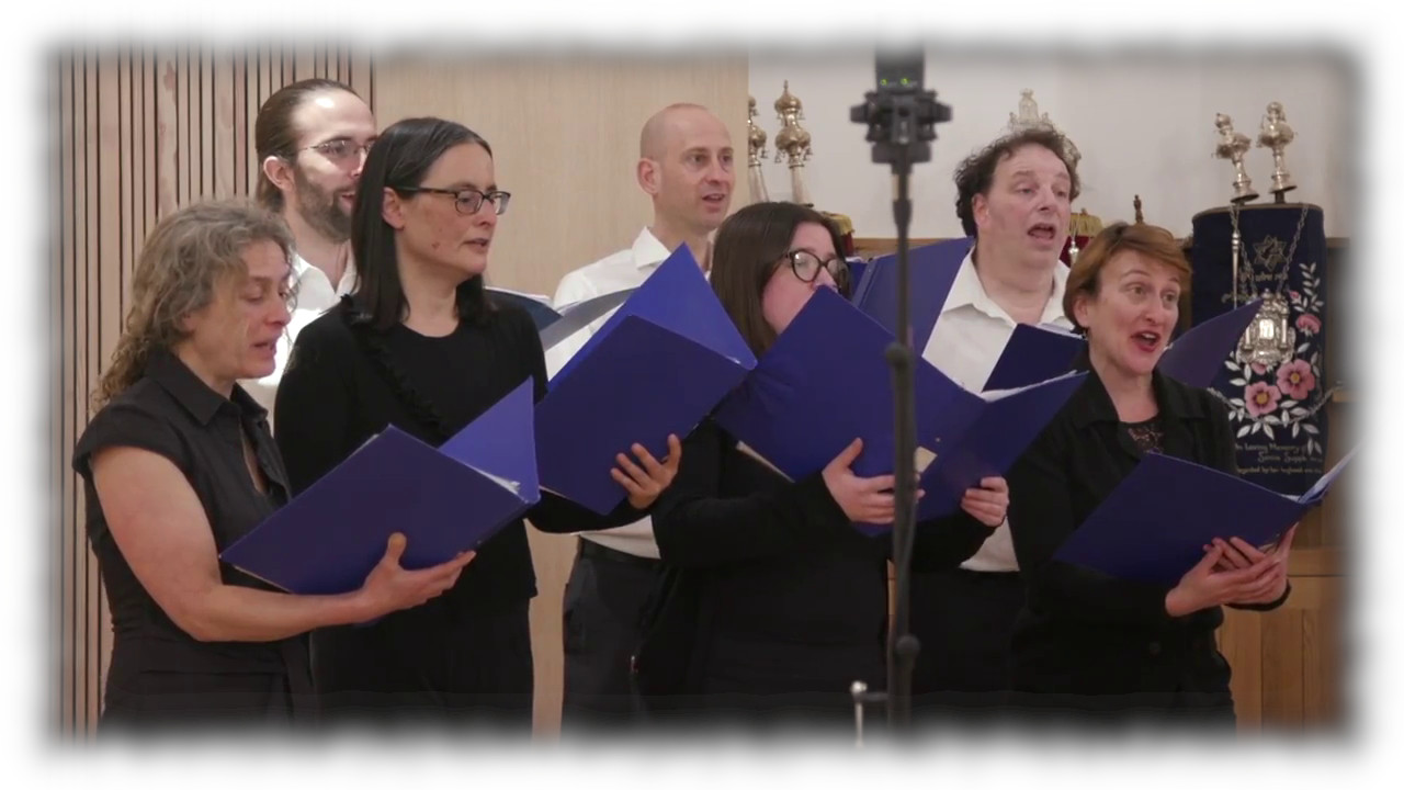 Kol Echad Choir group photo. Members standing next to a piano and singing joyfully.